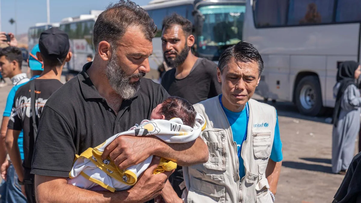 Ein Vater hält sein Baby im Arm. Neben ihm steht ein UNICEF-Helfer.